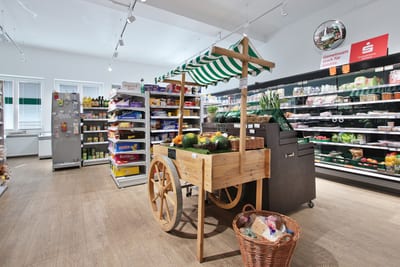 Verkaufsraum mit Obst und Gemüse auf einem Marktkarren aus Holz, dahinter Regale und Kühlregale