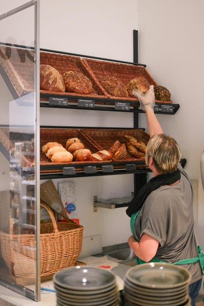 Mitarbeiterin mit grüner Schürze legt Brot in das Brotregal, darin Körbe mit Broten und Brötchen