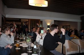 Gäste an einem langen Tisch heben die Hand, eine Frau hält eine grüne Karte hoch
