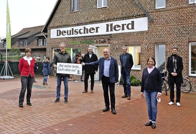 Neun Personen stehen mit Abstand vor dem Dorfladen „Deutscher Herd“, ein Mann hält ein Schild mit der Aufschrift „Wer weiter denkt, kauft näher ein!“