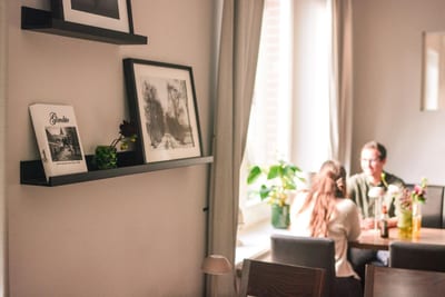Gäste an einem Tisch am Fenster im Dorfcafé, im Vordergrund ein Wandregal mit dem Buch „Gimbte“ und Schwarz-Weiß-Bildern