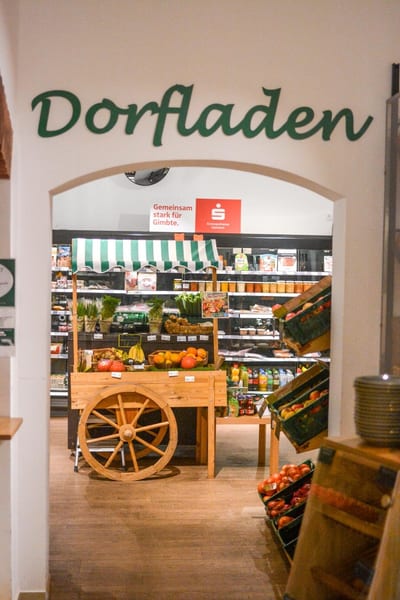 Eingang zum Dorfladen mit grünem Schriftzug über dem Torbogen, dahinter ein Marktkarren mit Obst und Gemüse und Kühlregale