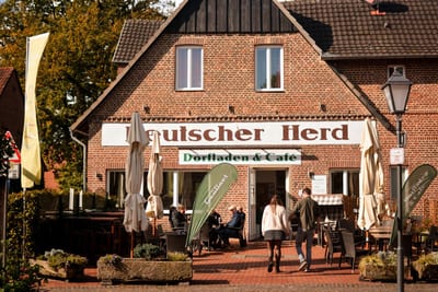 Backsteinhaus mit den Schriftzügen „Deutscher Herd“ und „Dorfladen & Café“, davor Gäste auf der Terrasse in der Sonne